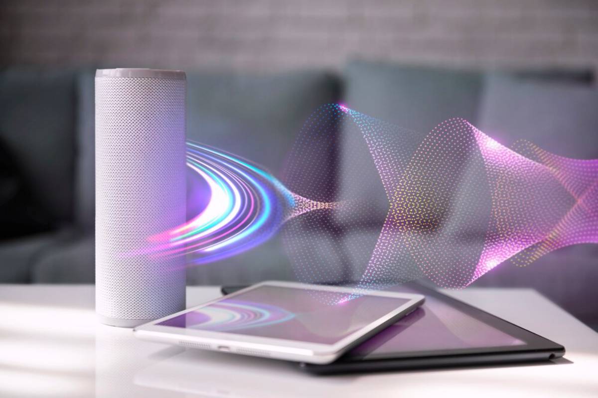 A white smart speaker emits colorful sound waves above a smartphone and tablet on a table, with a soft sofa in the background.