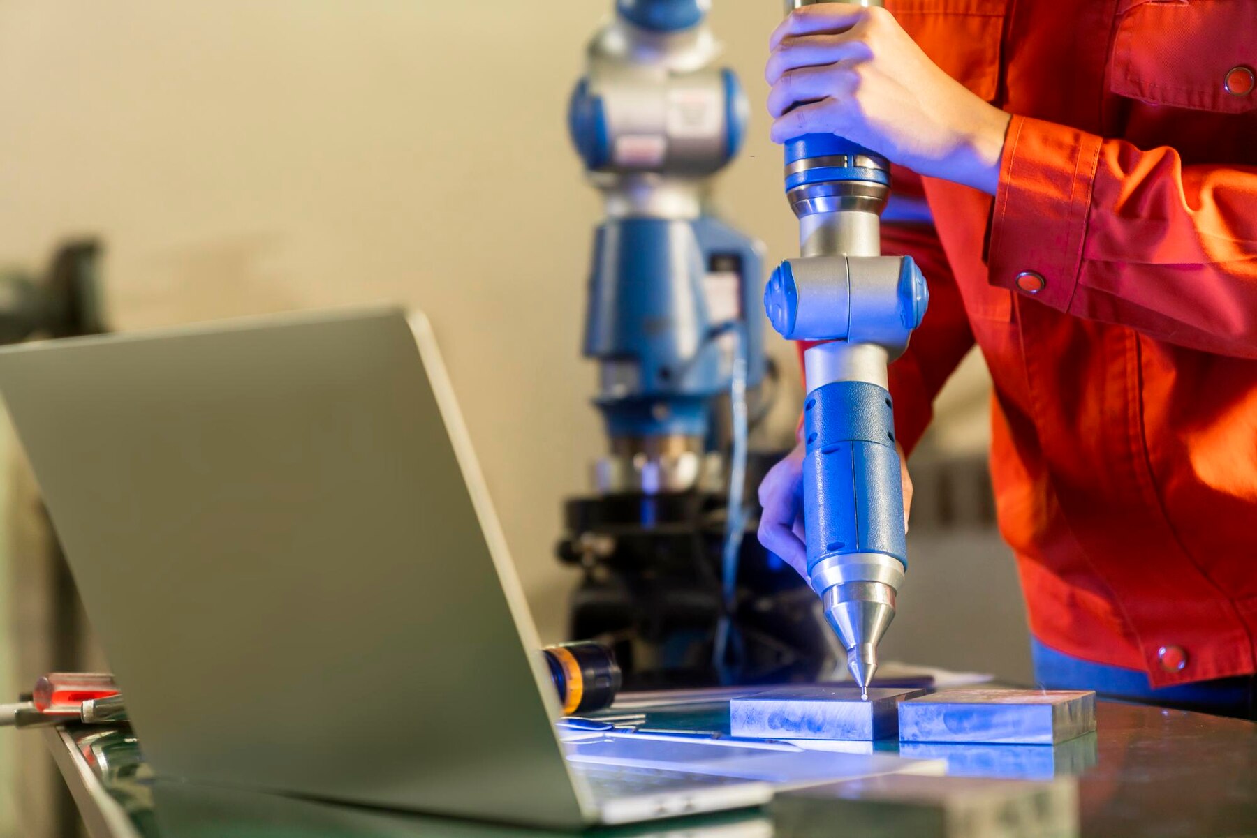 A technician in a red jacket uses a precision tool on metal pieces, with a laptop nearby in a workshop setting.