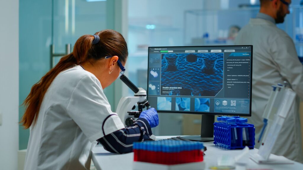 A researcher in a lab coat analyzes data from a microscope on a computer screen, surrounded by test tubes and lab equipment.