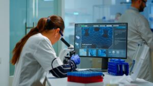 A researcher in a lab coat analyzes data from a microscope on a computer screen, surrounded by test tubes and lab equipment.