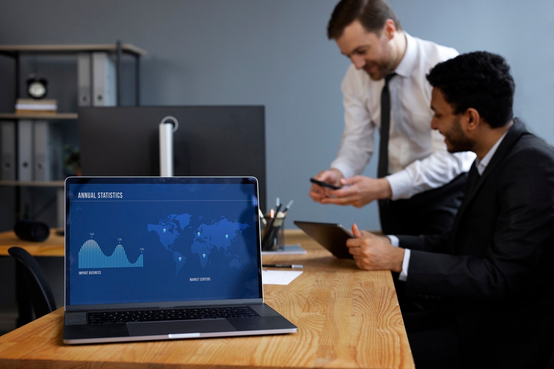 A modern office setting with a laptop displaying annual statistics and a world map, as two professionals engage in discussion.