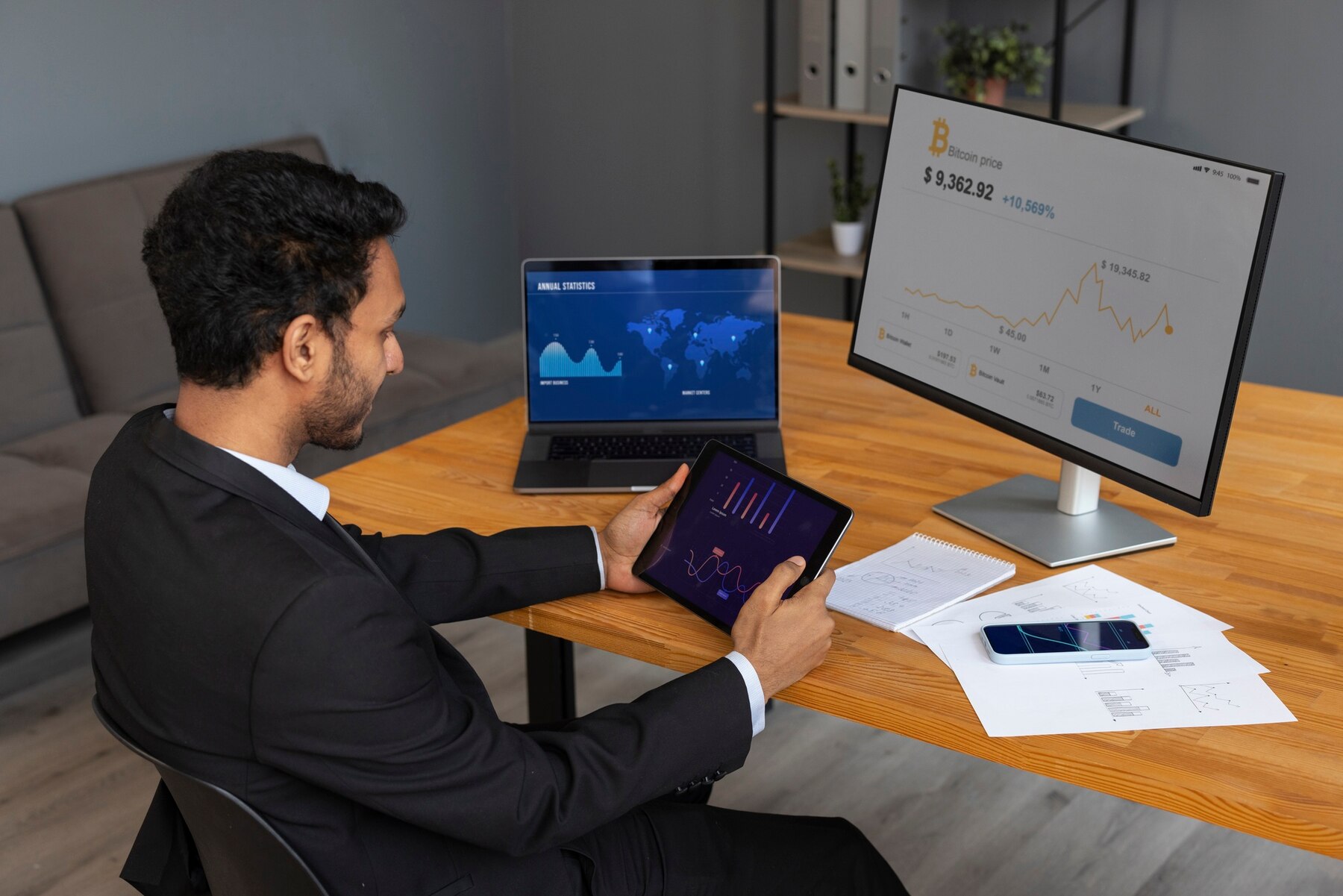 A businessman in a suit analyzes financial data on a tablet, laptop, and monitor, with reports and a smartphone on the desk.