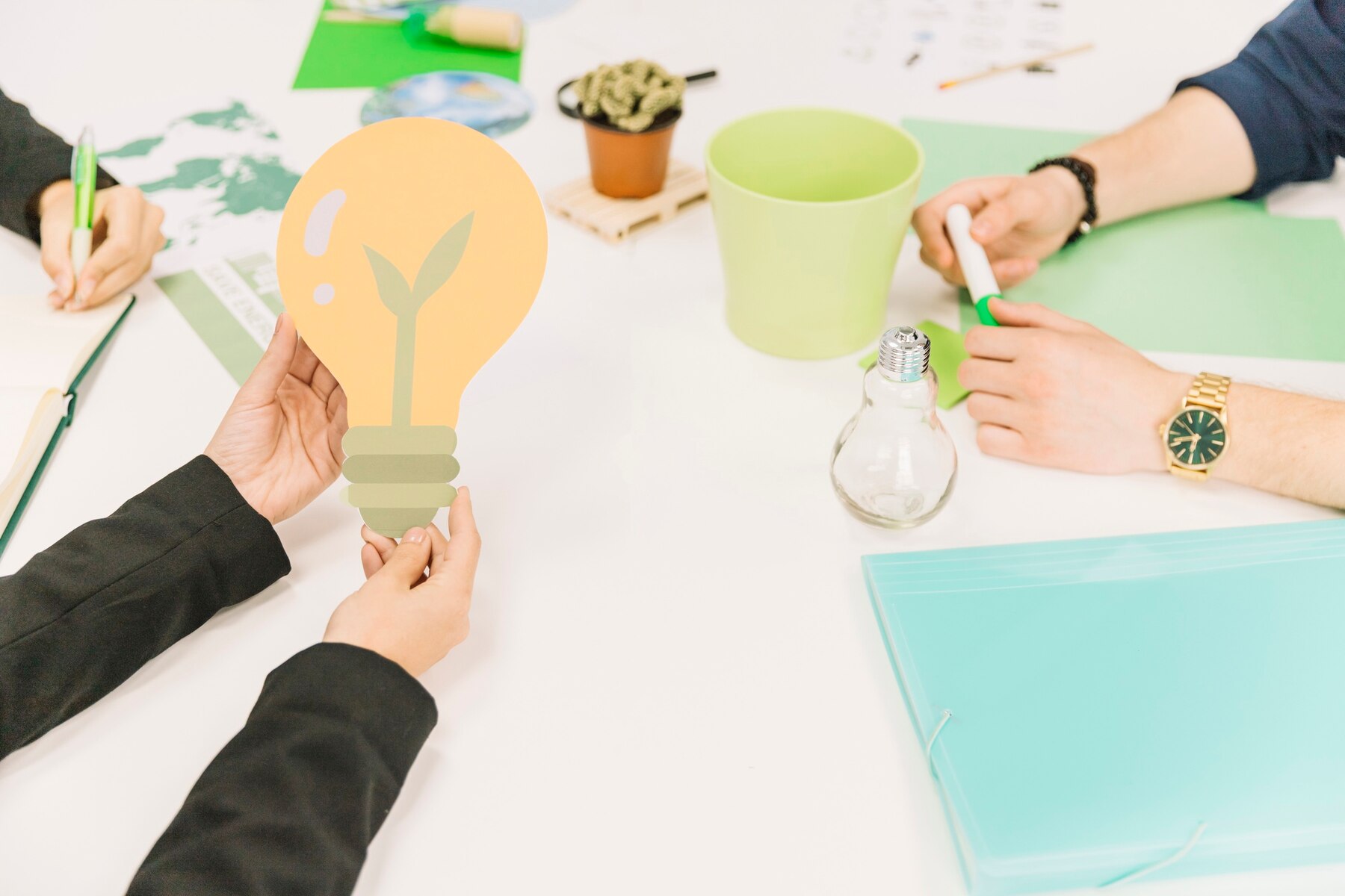 A person holds a green sprouting light bulb cutout at a workspace filled with plants, notebooks, and supplies, symbolizing eco-friendly ideas.