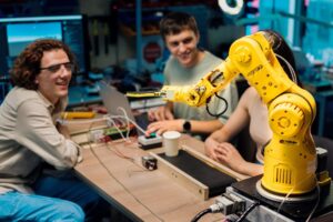A yellow robotic arm is positioned over a workbench filled with electronic equipment, while individuals engage in a discussion.