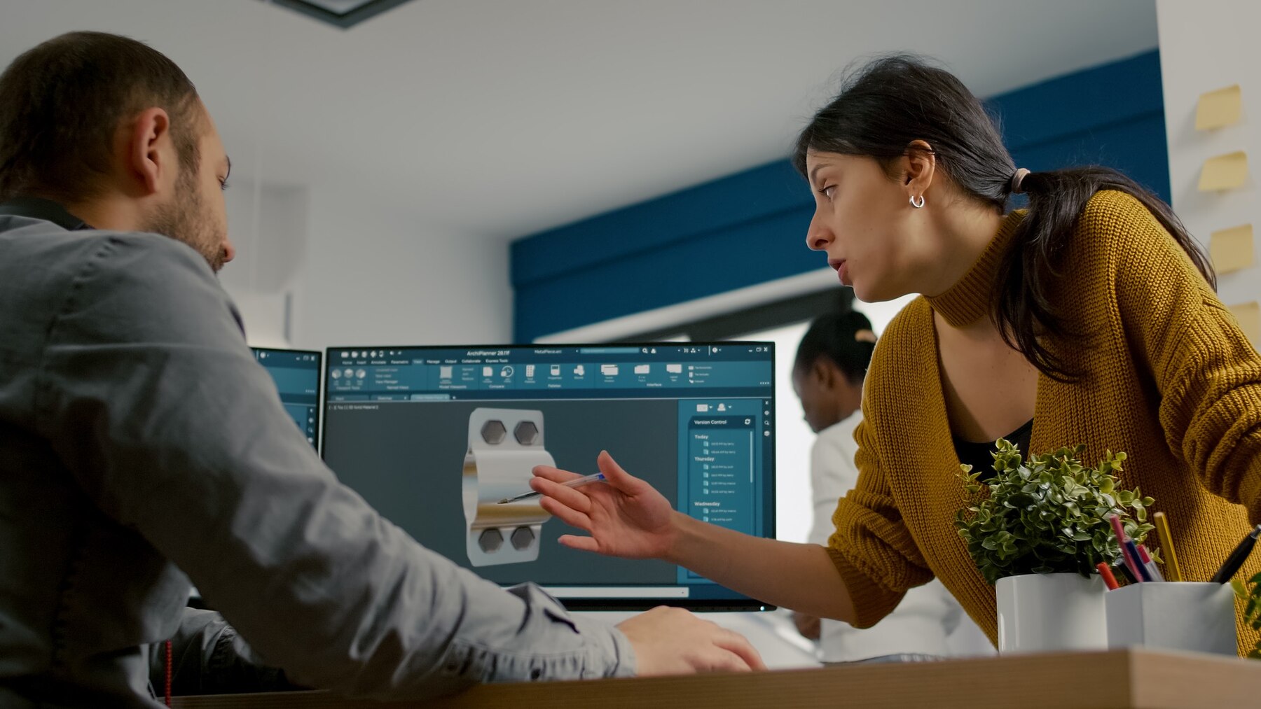 A woman in a mustard sweater gestures while discussing a design on a computer screen with a man in a gray shirt, plants on the desk.