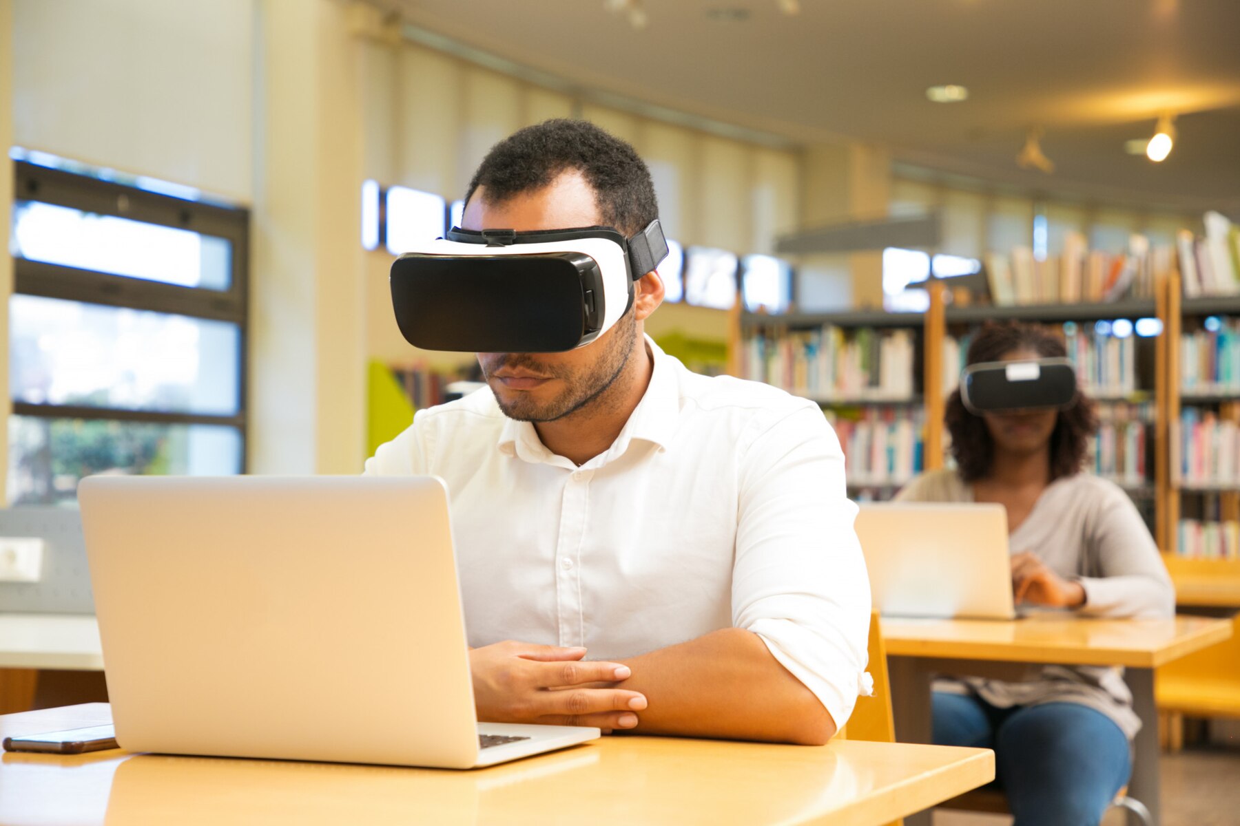 A student wearing a virtual reality headset sits at a wooden table in a library, focused on their laptop, surrounded by bookshelves.