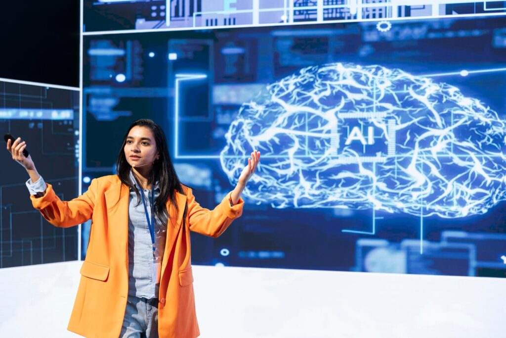 A speaker in an orange blazer gesturing while presenting on a digital display featuring a glowing brain and AI graphics.