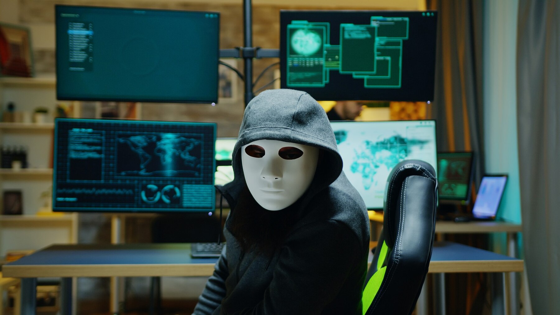 A person in a dark hoodie sits in front of multiple computer screens displaying green code and data analytics in a tech-filled room.