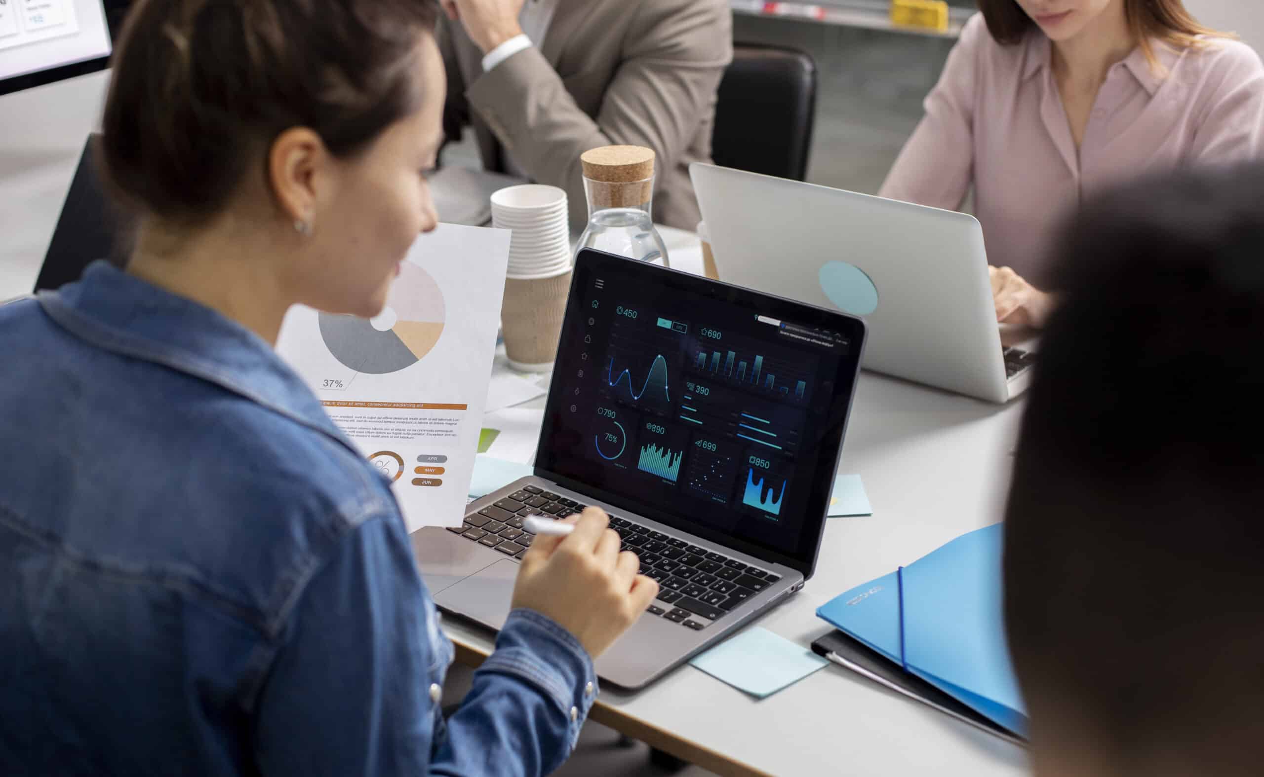 A person in a denim jacket analyzes data on a laptop, with graphs and charts on screen, surrounded by colleagues in a modern workspace.