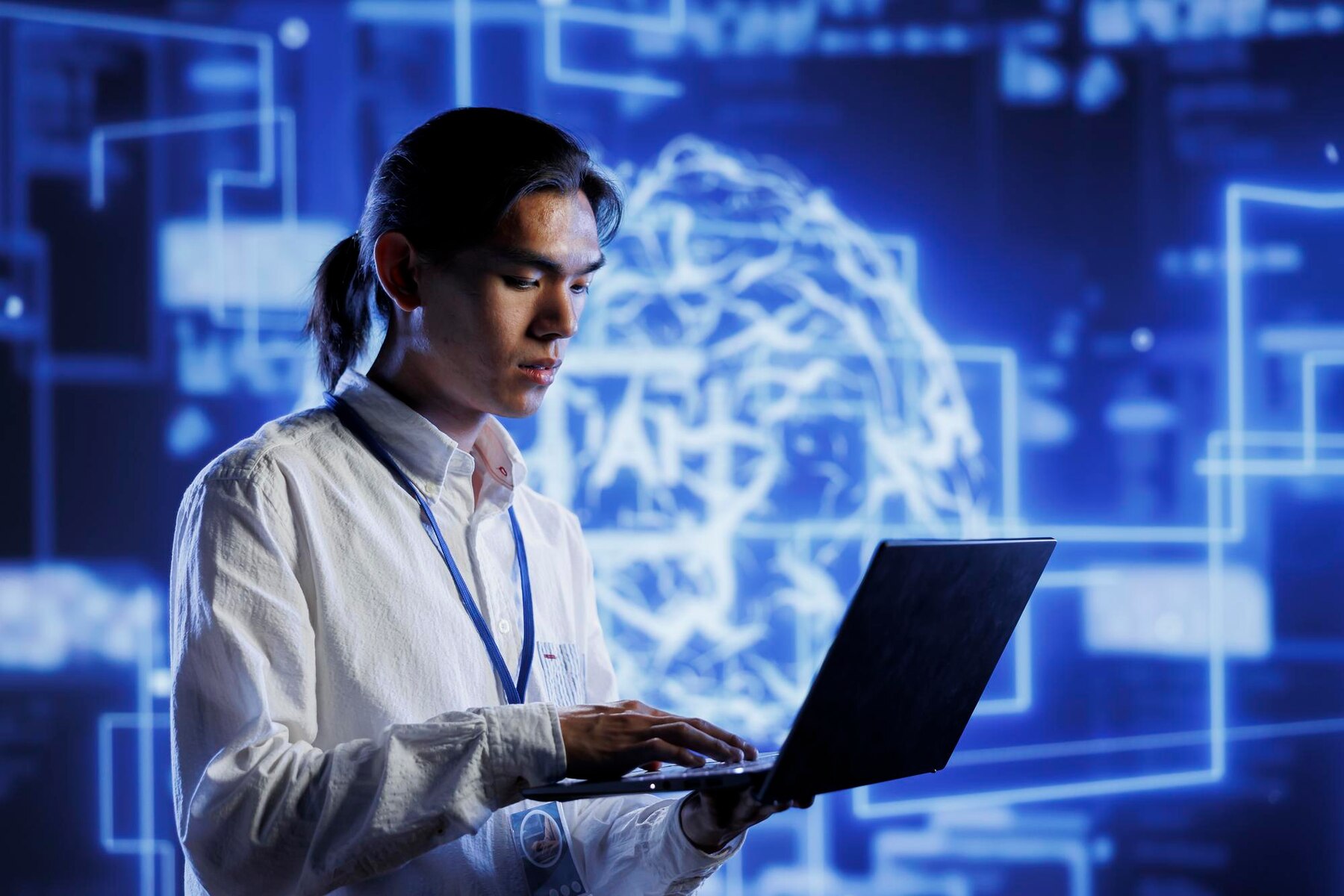 A person in a white shirt and lanyard types on a laptop, with a blue digital brain graphic in the background.