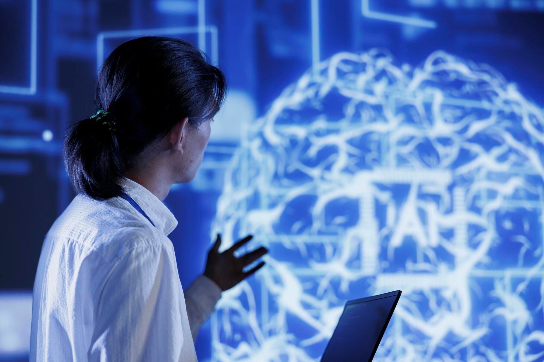 A person in a white shirt gestures towards a blue projection of a brain, while a laptop sits on the table in front of them.