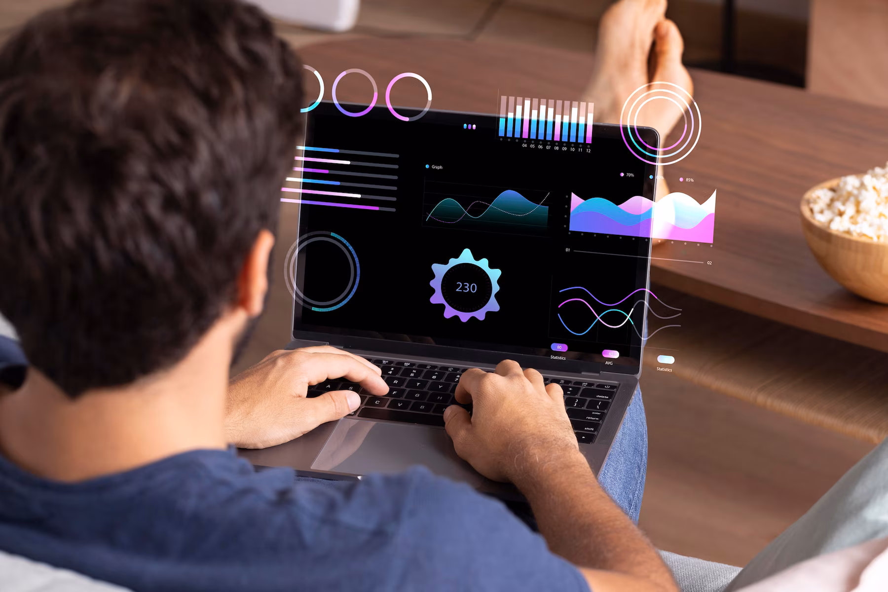 A man interacts with a laptop displaying colorful data visualizations and analytics while relaxing at home with popcorn nearby.