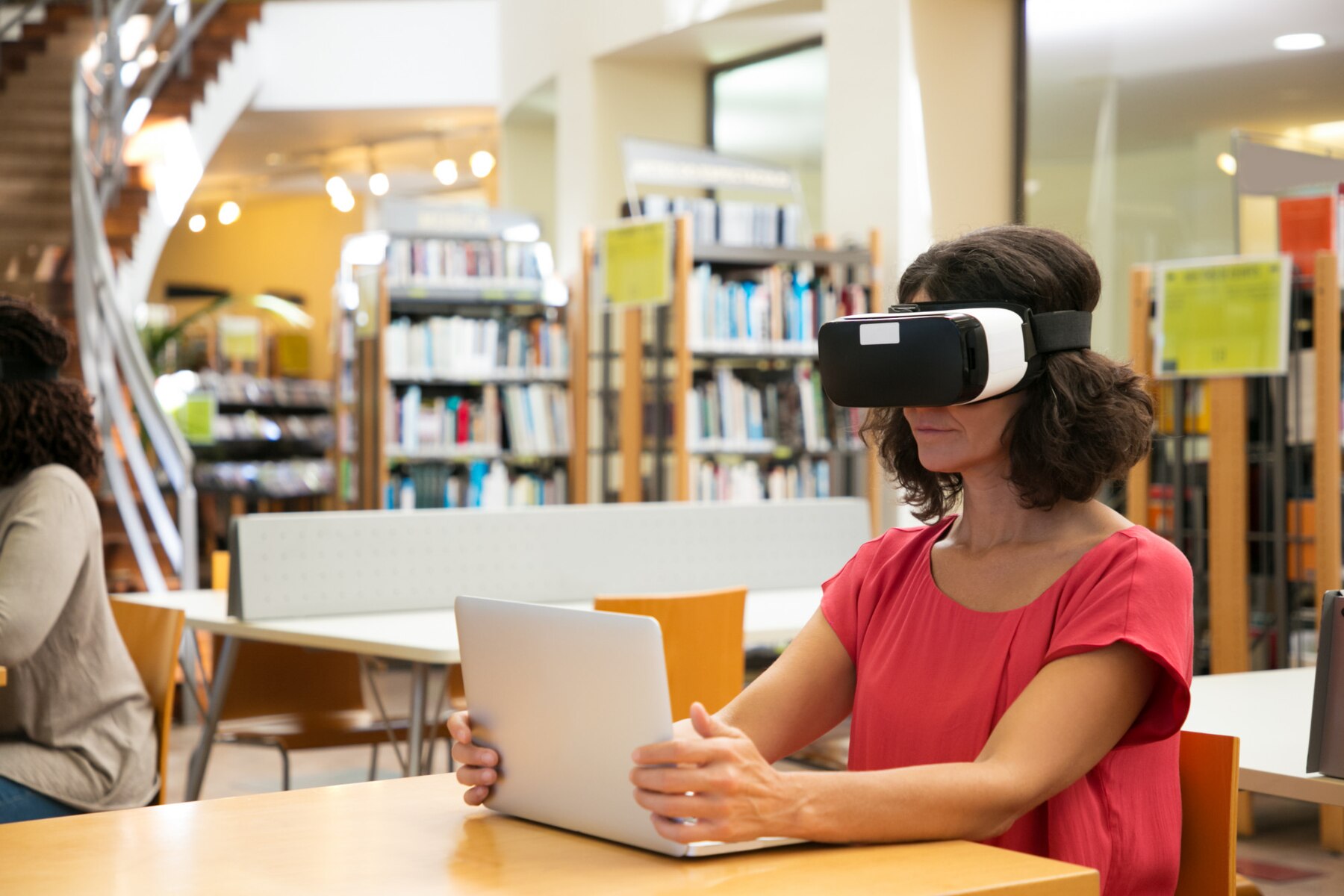 A person wearing a VR headset sits at a table in a library, using a laptop, surrounded by bookshelves in a bright space.