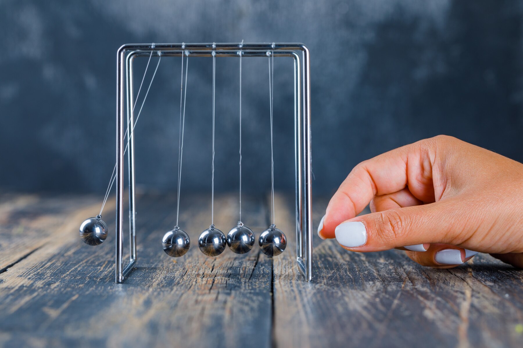 A hand is about to release a ball in a Newton's cradle, showcasing the principles of physics on a rustic wooden surface.