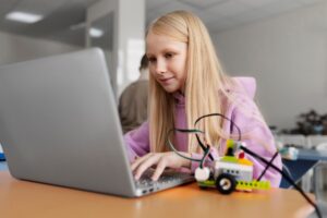 A young person in a pink hoodie typing on a laptop, with a colorful robotic toy beside them on a desk in a bright classroom.