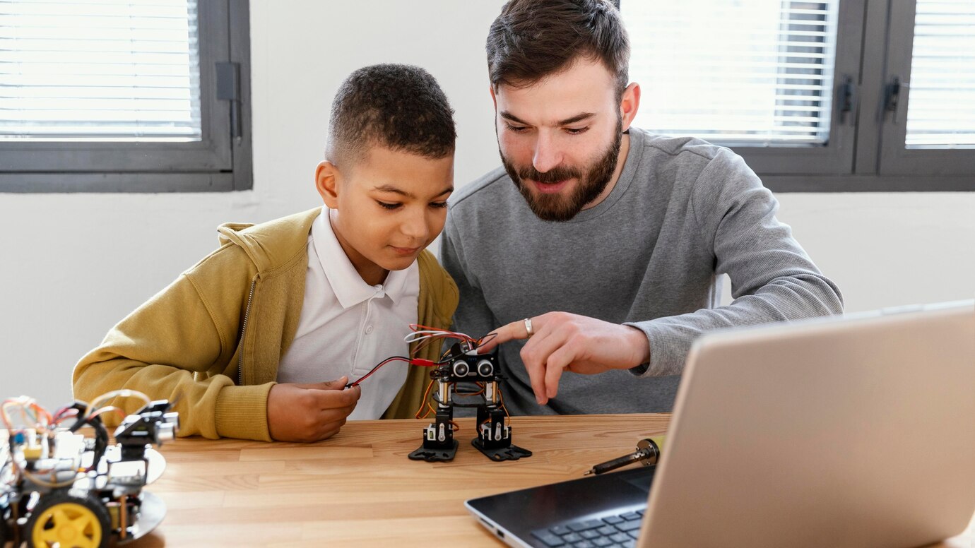 A child and an adult work together on a small robot, connecting wires over a wooden table with a laptop nearby.