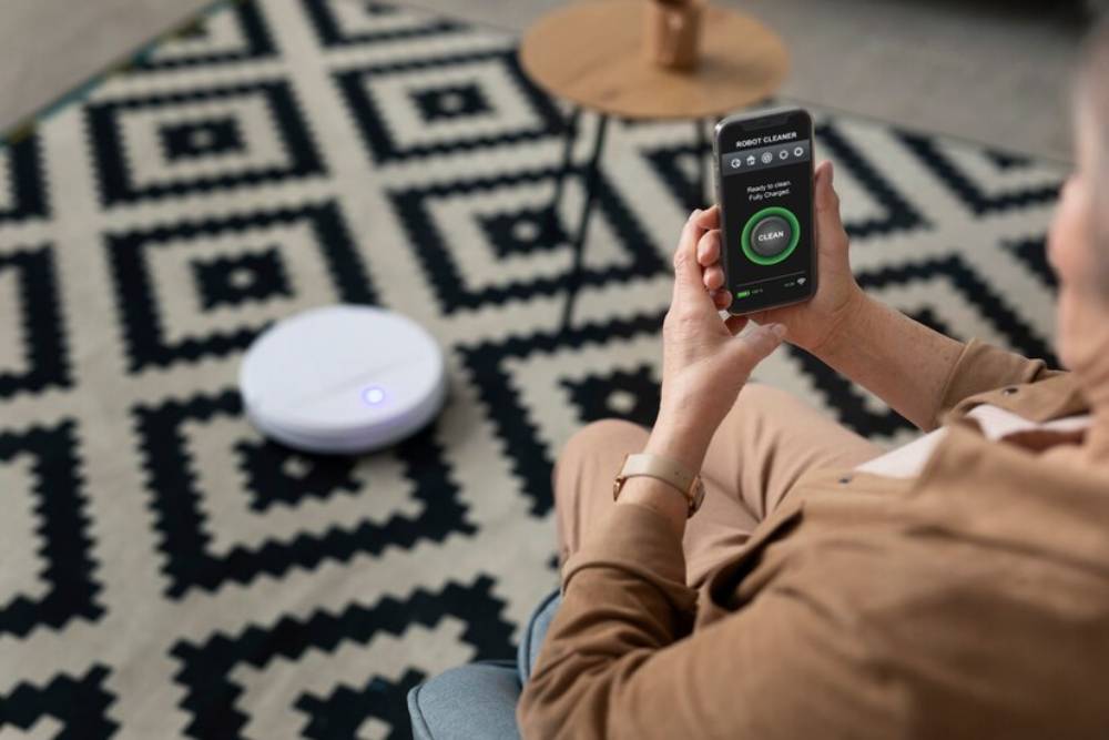 A person operates a robot vacuum through a smartphone app, sitting on a stylish floor rug with geometric patterns.