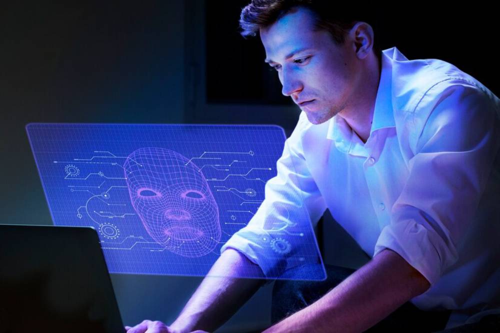 A person in a white shirt works on a laptop, interacting with a holographic display of a digital face and circuit patterns.