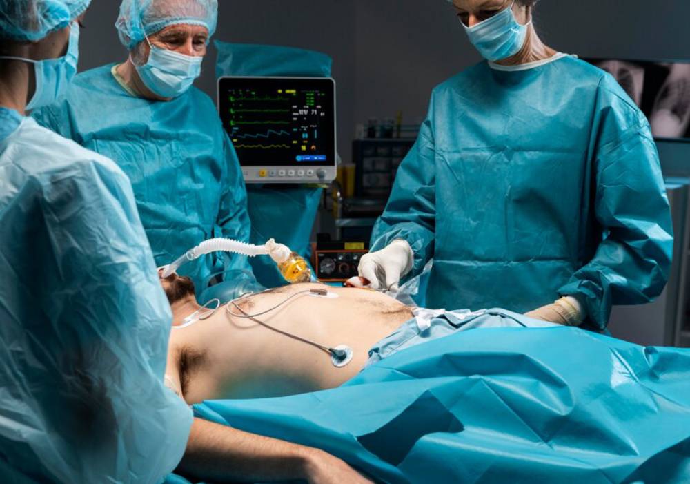 Surgical team in blue scrubs operates on a patient under anesthesia in an operating room. Medical monitor displays vital signs.