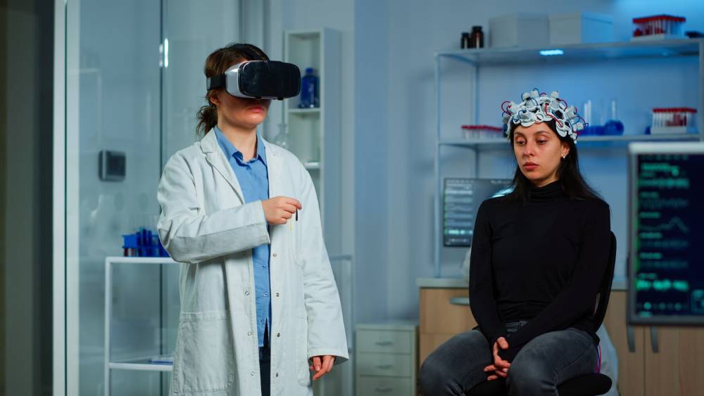 A scientist in a lab coat demonstrates virtual reality technology to a seated participant wearing a neuroimaging headset.