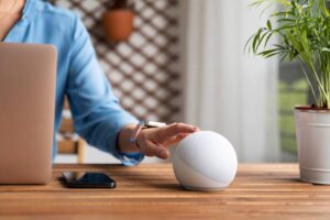 A person in a blue shirt touches a white smart speaker on a wooden desk, with a laptop and smartphone nearby, and a potted plant in the background.